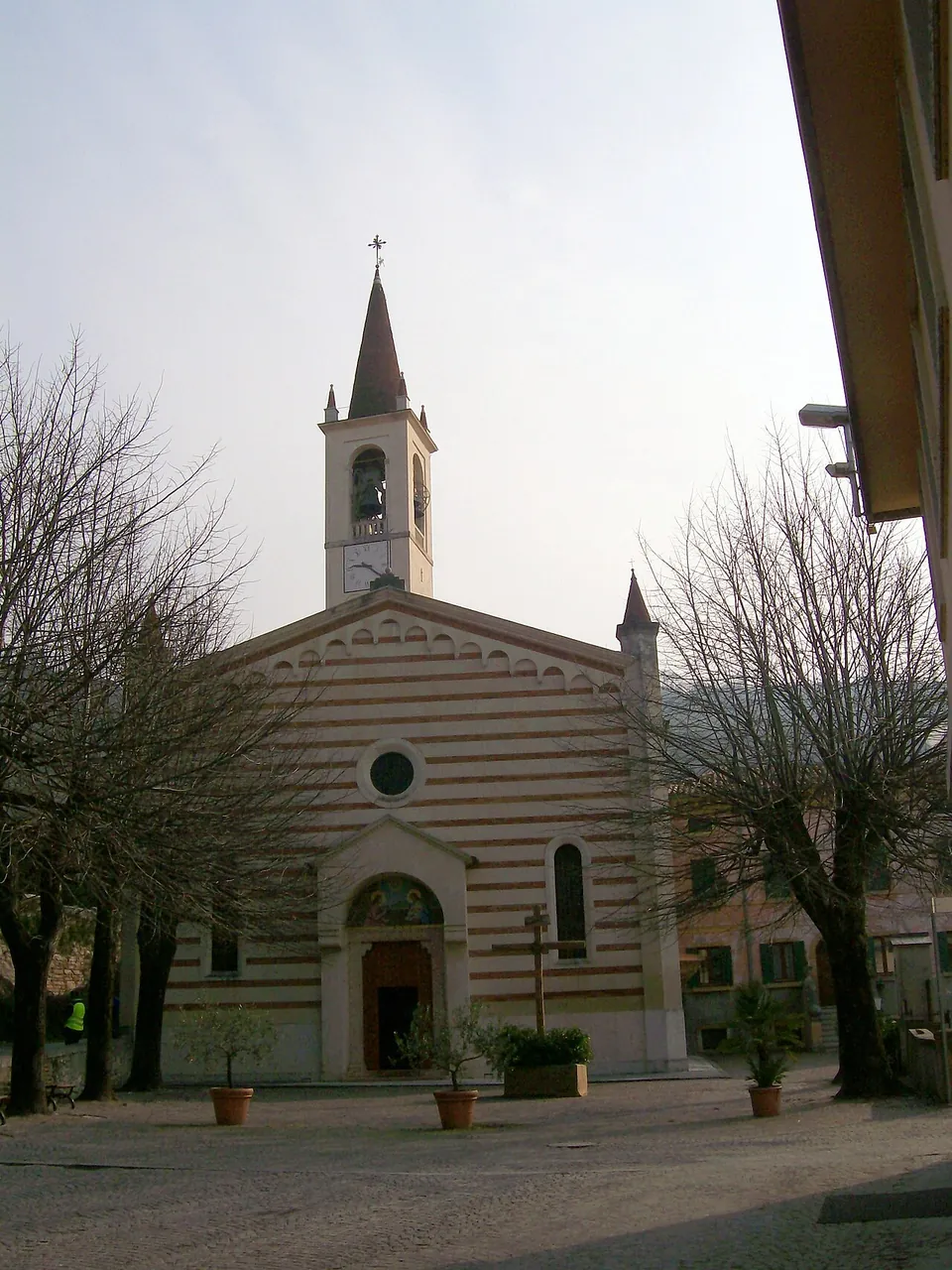 The front of a church with a striped gabled facade, a round window above an arched doorway, and a bell tower with a pointed spire behind, seen across a paved square with bare trees and potted plants.