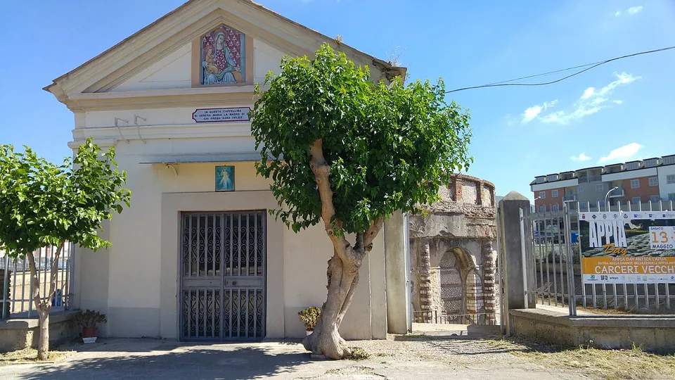 A small cream-painted chapel with a pediment, a holy image over the door and an iron gate, standing by the road; to its right, behind railings and a sign reading Carceri Vecchie, part of the tomb's brick-banded wall shows.