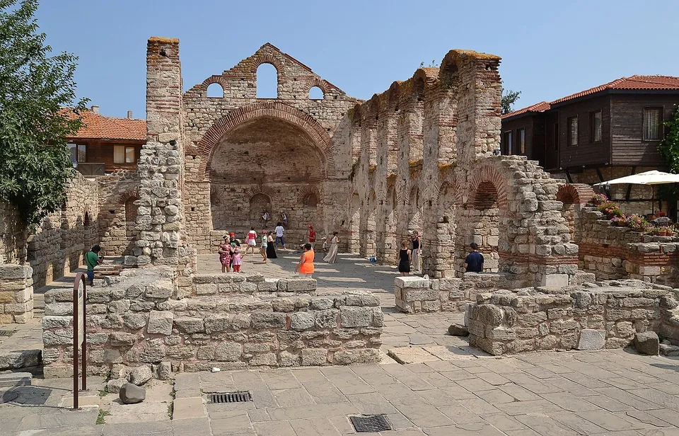 The roofless shell of a large stone and brick basilica, its apse wall with an arch and three windows and a row of arches along one side, with tourists walking inside and old houses around it.