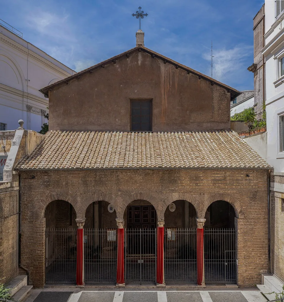 A plain brick church front with a tiled roof sloping over a portico of five round arches on red columns, closed with iron grilles, on a paved forecourt.