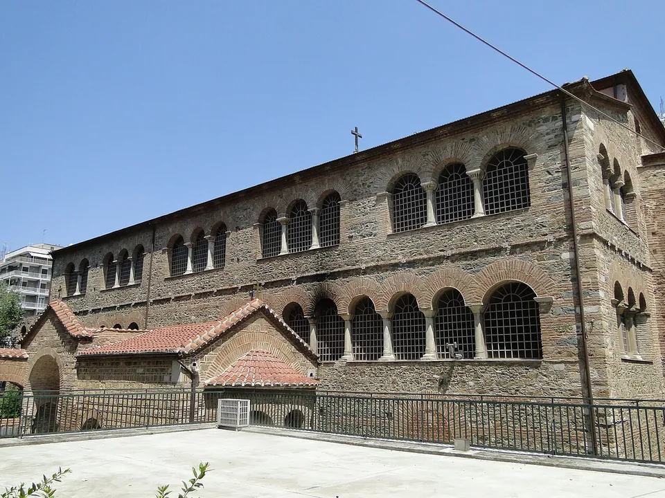 The long side of a large basilica of stone and brick, with two tiers of arched windows divided by slender columns, a lower tiled porch in front and modern apartment blocks behind.