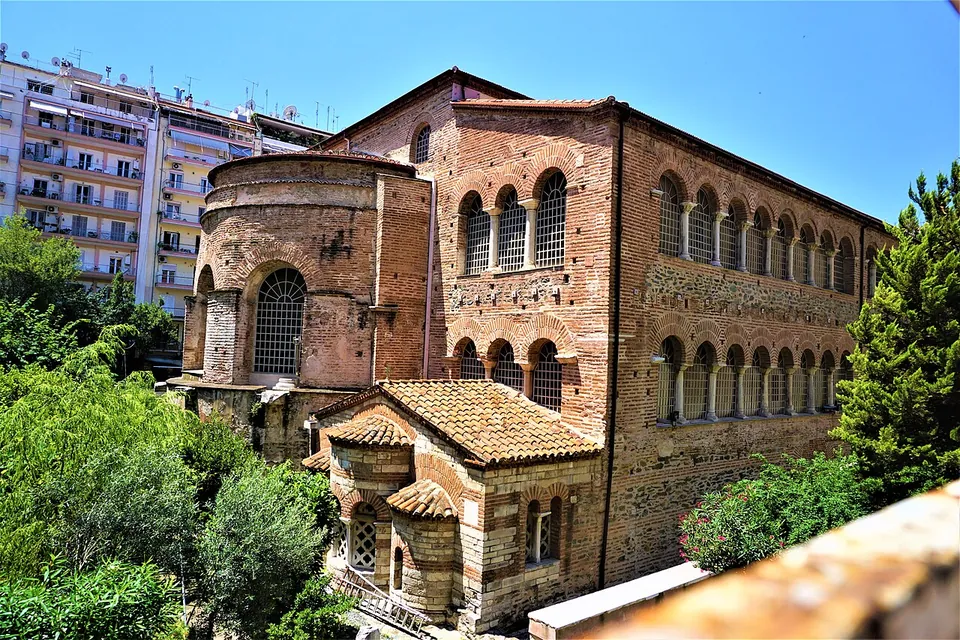 A basilica of red brick and stone with rows of arched windows and a round-ended annex, seen from above over green trees, with apartment blocks behind.