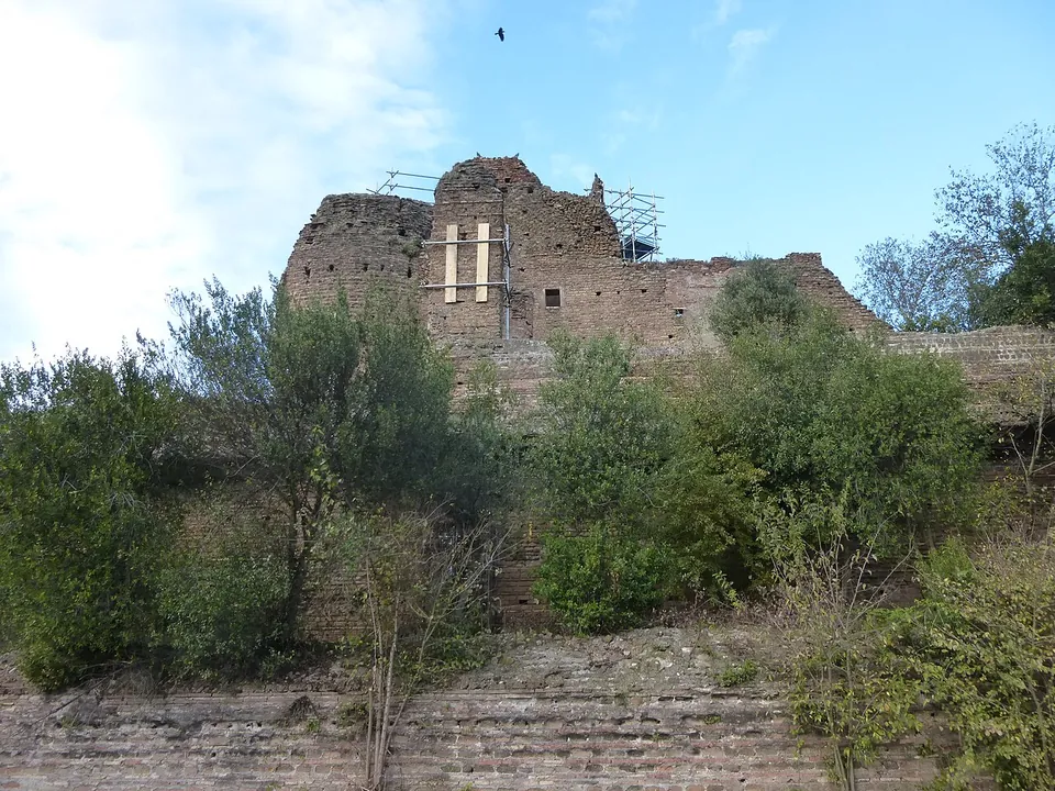 A tall ruined block of brick, braced with timber on its face and topped by scaffolding, rises above trees and shrubs, with a low brick wall in the foreground.