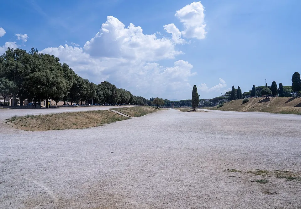 A broad, pale gravel track running straight away from the camera between low grassy banks, umbrella pines along the left side and cypresses on the right, under a blue sky with large white clouds.