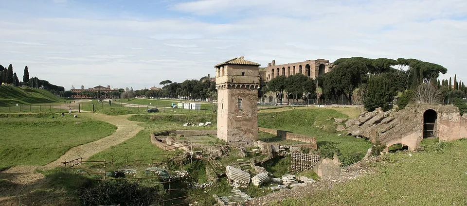 Excavated Roman walls and scattered blocks of stone in a fenced, grassy hollow, a square medieval tower with a tiled roof rising in the middle, and tall brick ruins above the trees behind.