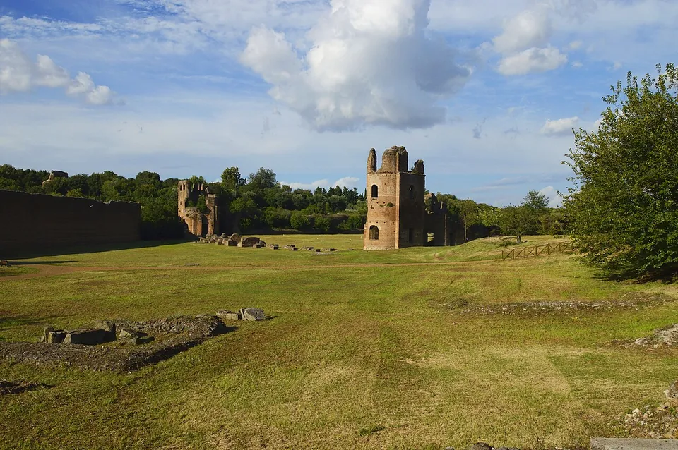 A long, flat grassy arena with scattered stone blocks, bounded on the left by a straight brick wall; at the far end stand two ruined brick towers, the nearer one tall with arched openings, under a blue sky with white clouds.