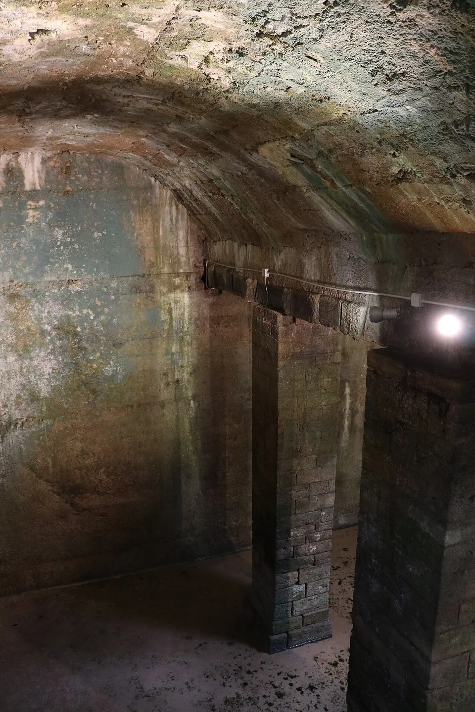 A dim underground chamber with a vaulted ceiling carried on square masonry piers, lit by a single lamp.