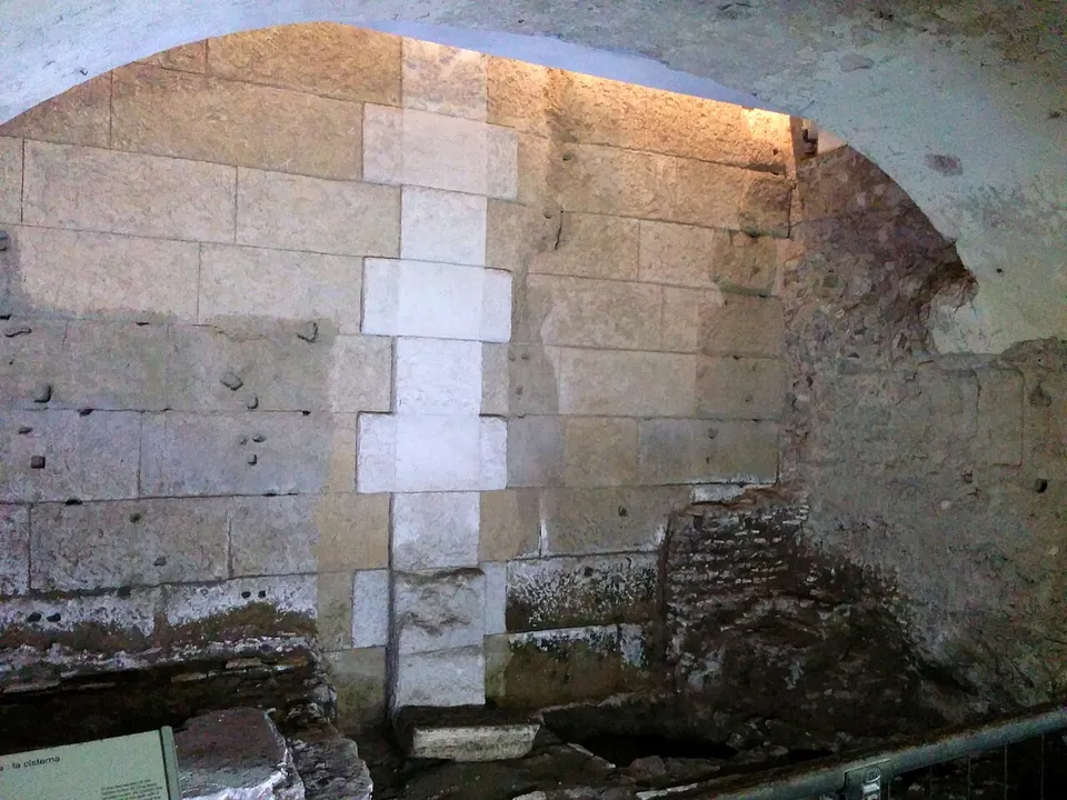 A lit wall of large squared stone blocks under an arch, with a vertical strip of paler blocks set into it, brickwork low down at the right, and an information panel and a railing at its foot.