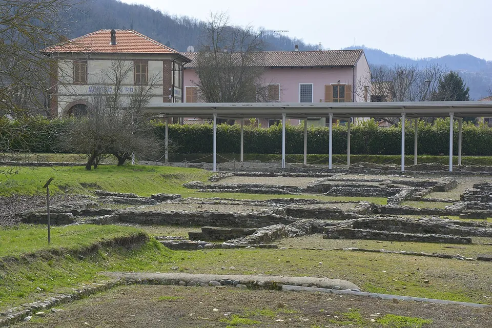 A spread of low stone foundation walls in grass and bare earth, with a long open-sided canopy on thin white posts behind them and a house and wooded hills in the background.