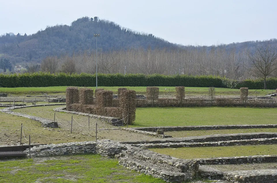 Low walls of rounded river stones and rubble outlining rooms in a mown lawn, behind them a row of tall, square-clipped hedge blocks in winter brown, a long green hedge and a wooded hill beyond.