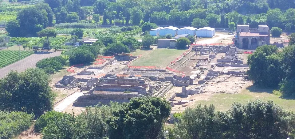 A view from high above over the excavated lower city, a grid of low walls and cleared ground with white site sheds, framed by woodland and farmland, a farmhouse at the far edge.