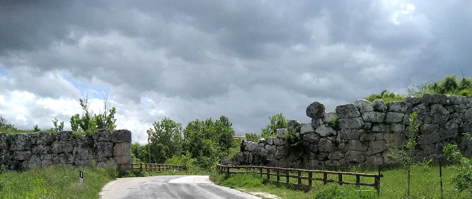 A narrow road passing through a gap in a high wall of huge irregular stone blocks fitted closely together, with a wooden fence along the verge and a stormy sky.