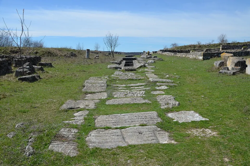 Large flat paving slabs set in the grass in a line, leading between low ruined walls towards a single standing column fragment.