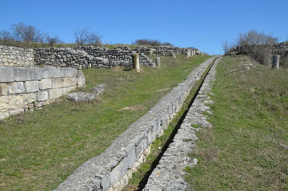 A grassy street running between walls of squared stone blocks, with a narrow stone-lined channel down its middle and a few broken column shafts standing beyond.