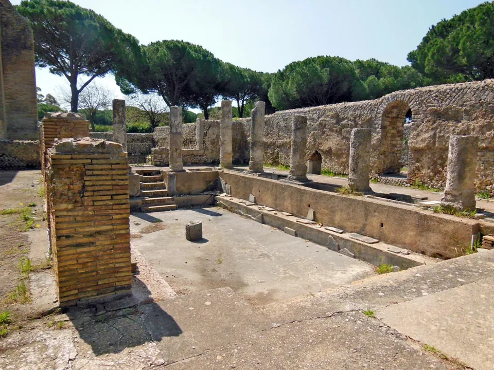 A long rectangular sunken basin with a stepped edge, lined along one side by a row of broken column shafts, with a high stone wall pierced by an arched opening behind and umbrella pines above.