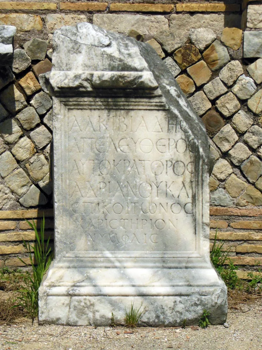 A white marble altar with a moulded top and base, its face cut with several lines of Greek capital letters, standing against a wall of squared stone blocks.