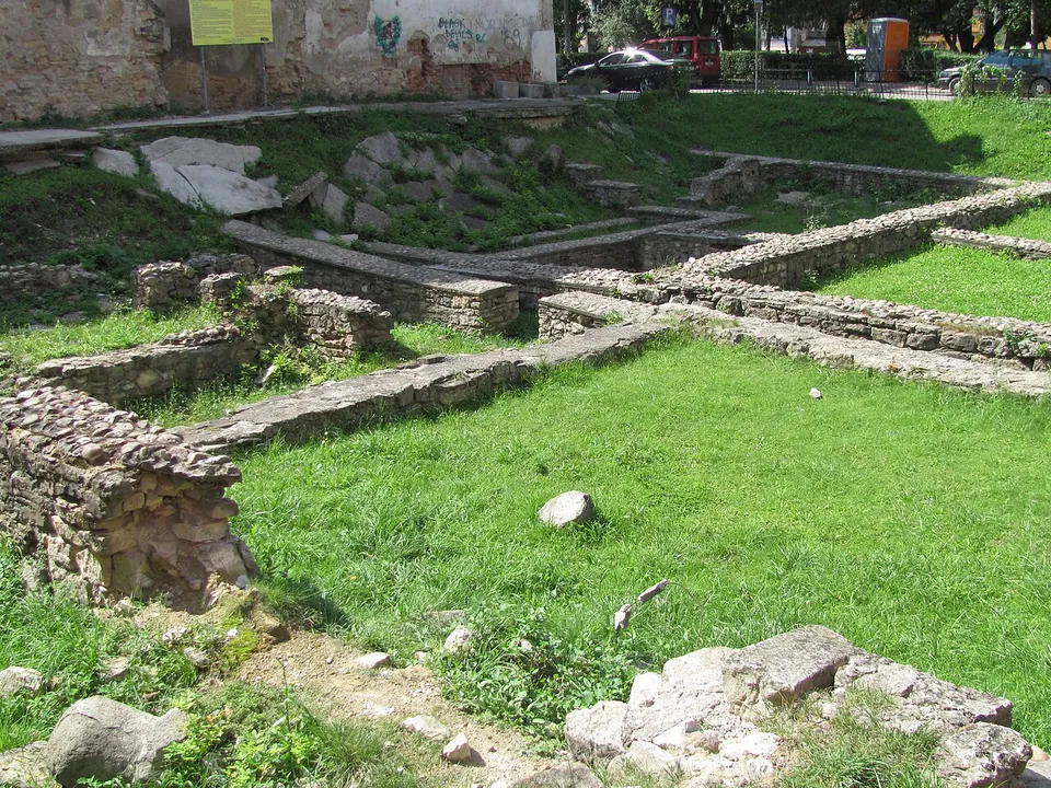 Low walls of mortared stone outlining rooms in a mown lawn below street level, with a ruined old wall, parked cars and a street behind.