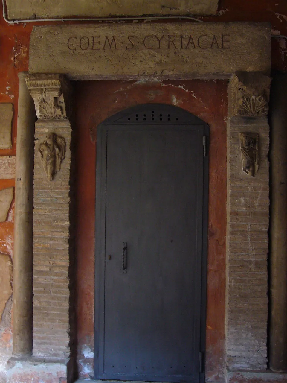 A plain grey metal door in a brick doorway framed by two small carved stone capitals and a stone lintel lettered COEM S CYRIACAE, set in a red-plastered wall.