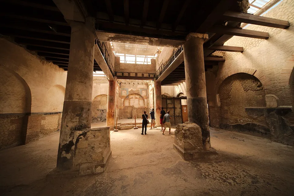 A dim interior hall under a timber ceiling, with two thick plastered columns in the foreground, a skylight in the roof and a painted shrine with a doorway at the far end; a few visitors stand inside.