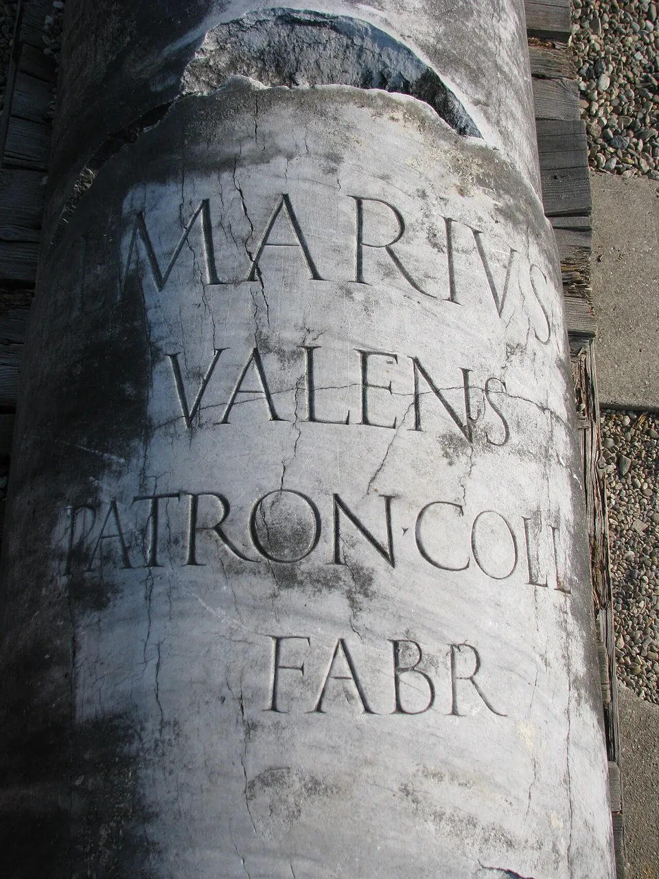 Close-up of a weathered white stone column carved with large Roman capitals reading MARIVS, VALENS, PATRON COLL, FABR.