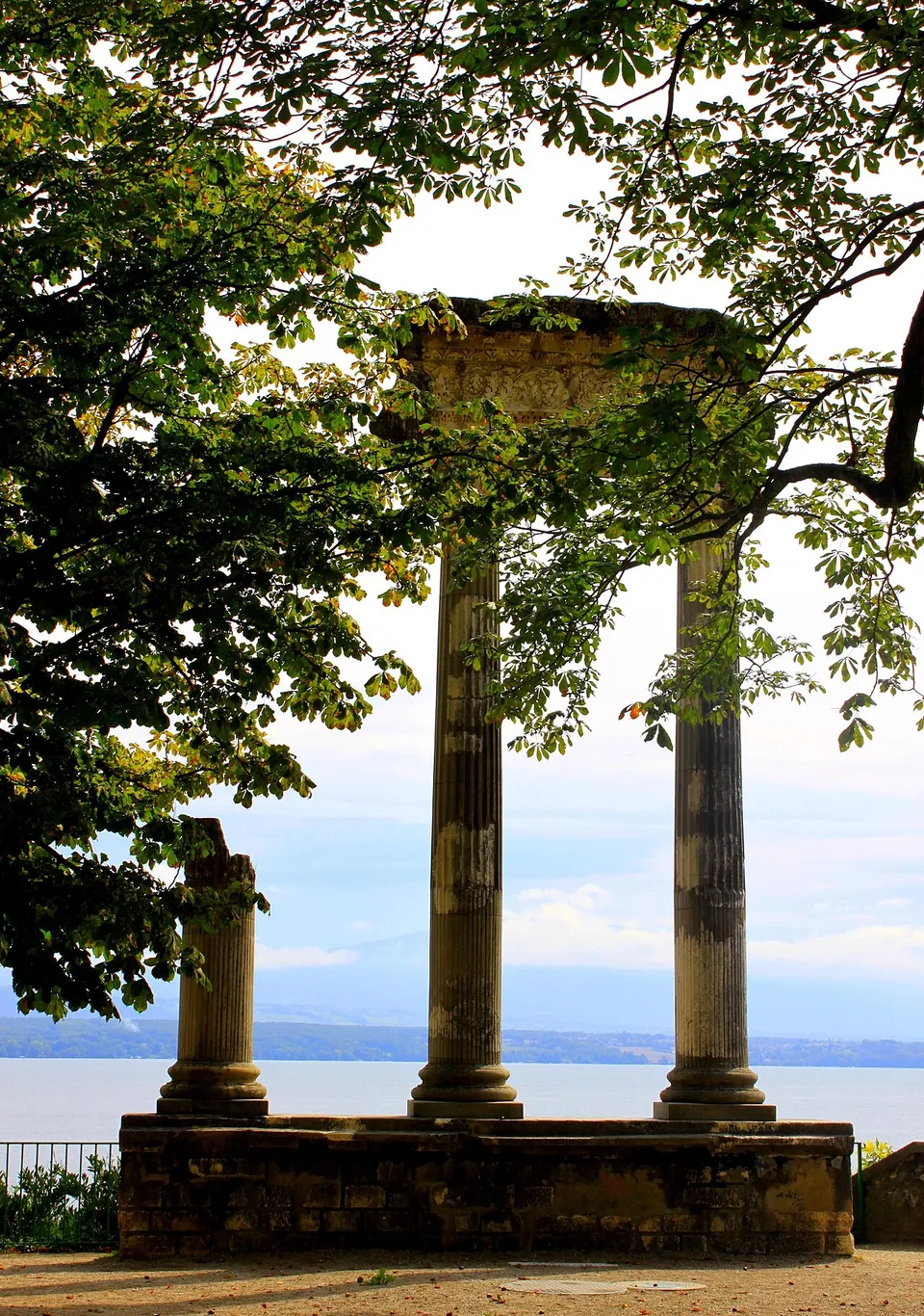 Three fluted stone columns on a low stone platform under chestnut branches, two tall ones carrying a weathered stone block across their tops and a shorter broken one beside them, with the lake and distant hills behind.