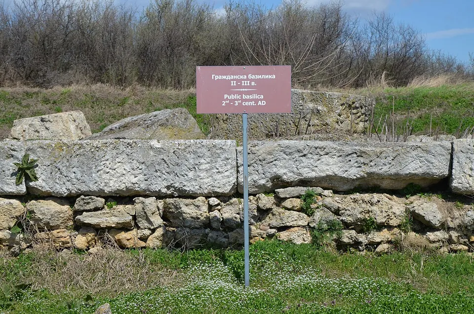 A small maroon sign on a post reading "Public basilica, 2nd–3rd cent. AD" in Bulgarian and English, in front of a low wall of large squared stone blocks set on rubble masonry.