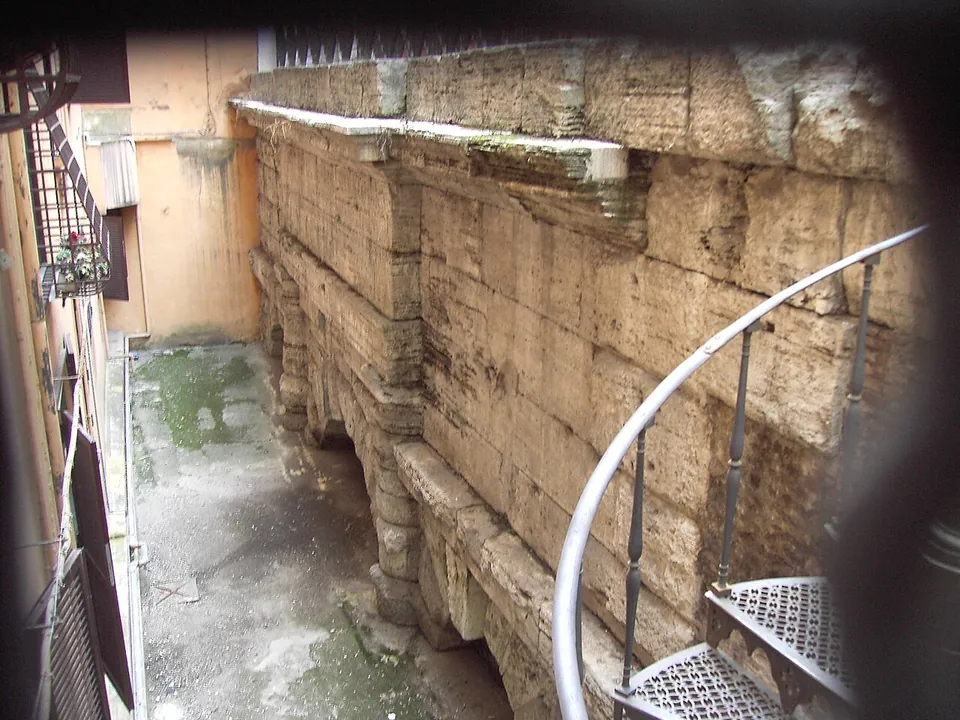 Seen from above through a railing, a long wall of massive weathered stone blocks with the tops of arches at its foot stands in a narrow sunken yard between house walls, a spiral iron staircase leading down in the foreground.