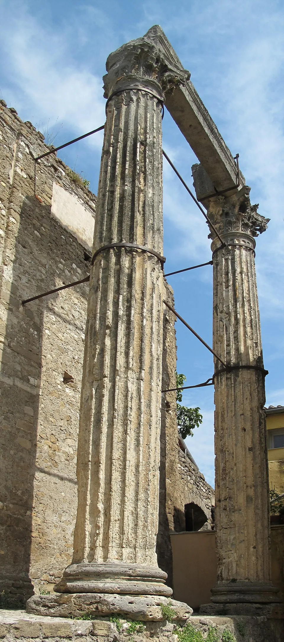Two tall fluted stone columns carrying a section of stone beam, held to a neighbouring wall by iron ties, against a blue sky.