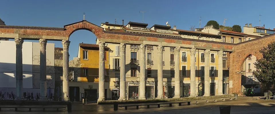 A long row of tall fluted columns with Corinthian capitals carrying a stretch of entablature, closed at both ends by brick arches, standing along a paved square in front of a terrace of pale town houses.