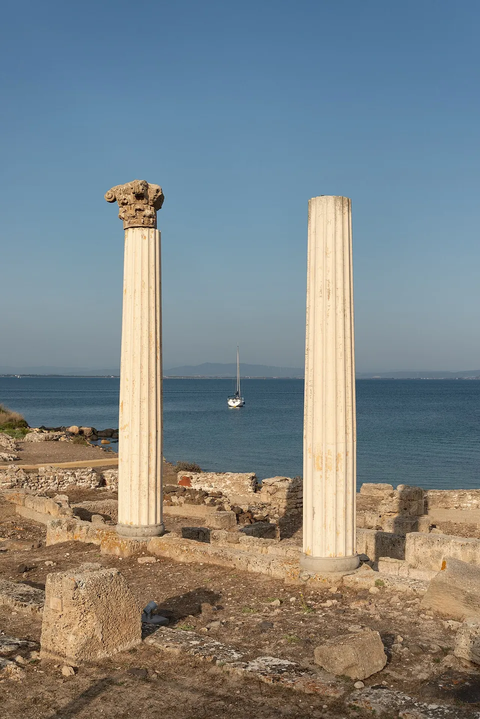 Two fluted white columns stand among low stone walls, the left one topped by a Corinthian capital, with calm sea and a moored sailing boat behind them.