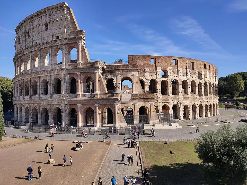 The Colosseum in sunshine seen from raised ground. On the left the outer wall rises in three tiers of arches with a solid top storey; on the right it breaks off to a lower ring of arches. Visitors cross the paved square in front.