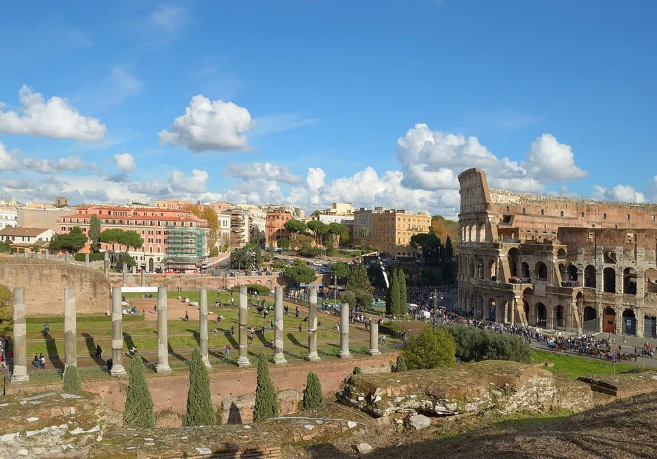The Colosseum's tiers of arches under a blue sky with white clouds, seen from raised ground where rows of column shafts stand on a lawn, with modern apartment blocks to the left.