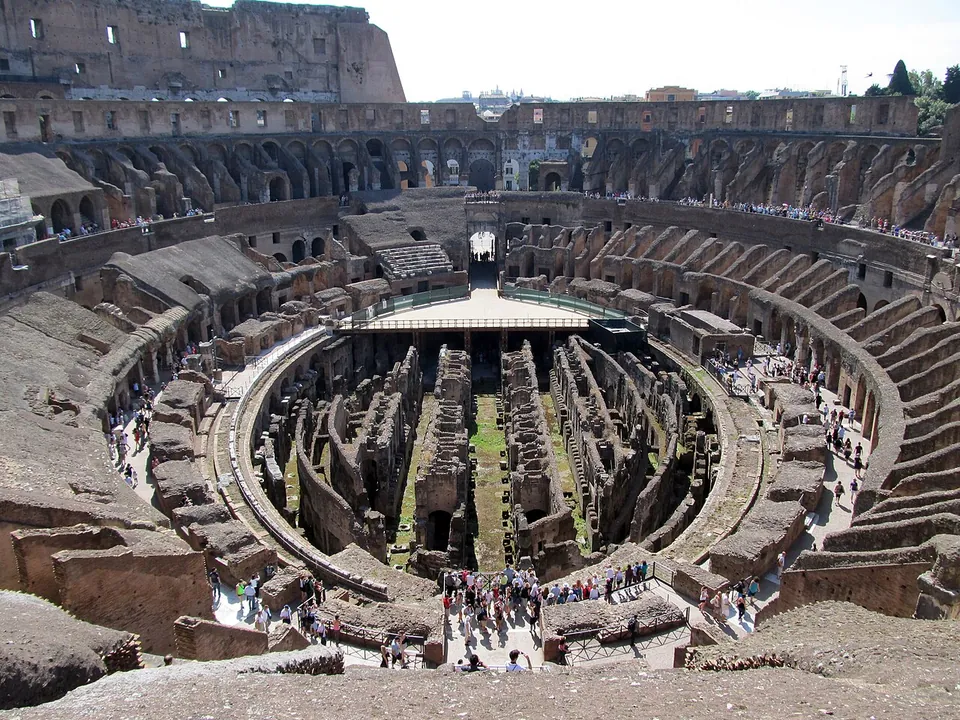 The inside of the amphitheatre from its top level. Ruined tiers of brick and stone run round an oval; part of the arena floor is laid at the far end, and in the middle the brick walls and passages of the basement lie open, with visitors along the edges.