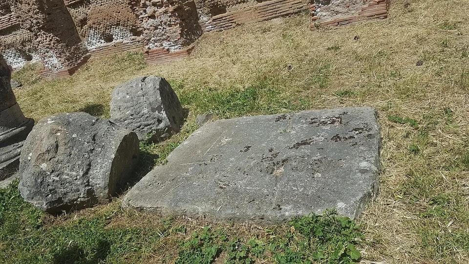 Two fallen column drums and a large flat stone block lying in the grass in front of ruined brick walls.