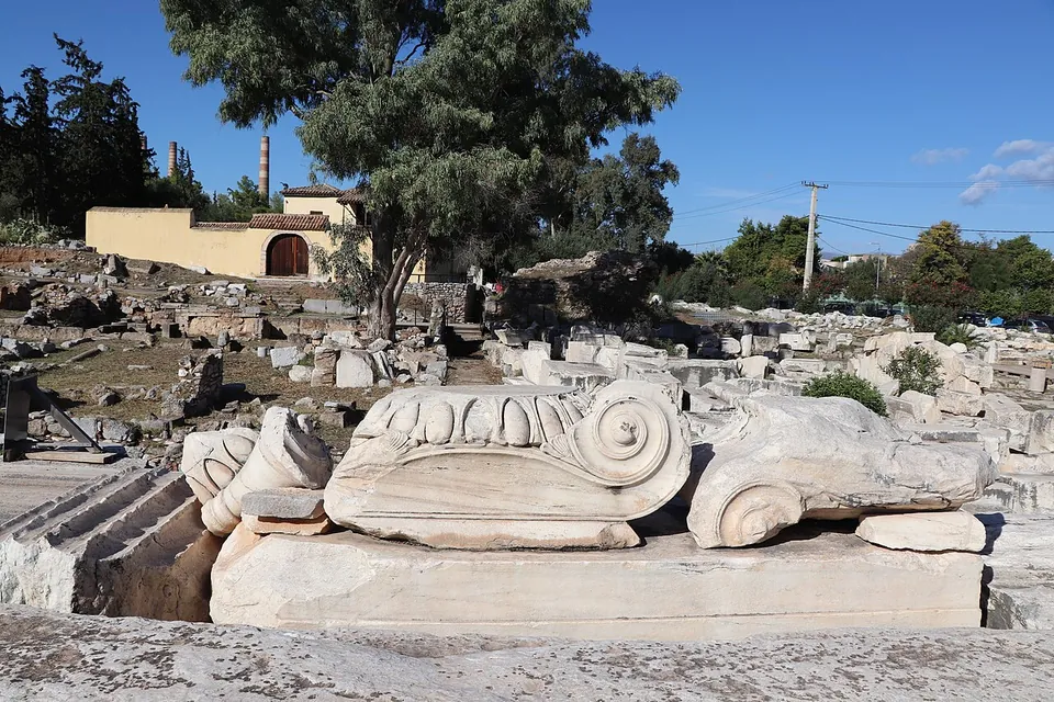 Large carved marble capitals with spiral volutes and egg-and-dart moulding laid on blocks in the sun, with ruins, trees and a house behind.