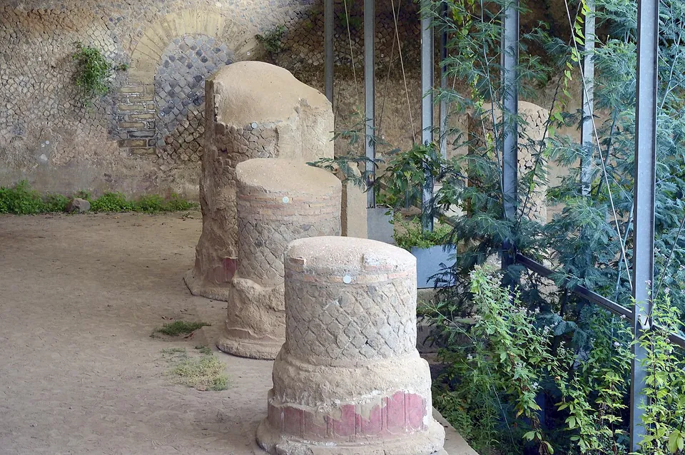 Three broken column stumps in a row, their surfaces laid in a net pattern of small stones with a band of red at the base, in front of a wall faced with small square stones set diagonally.