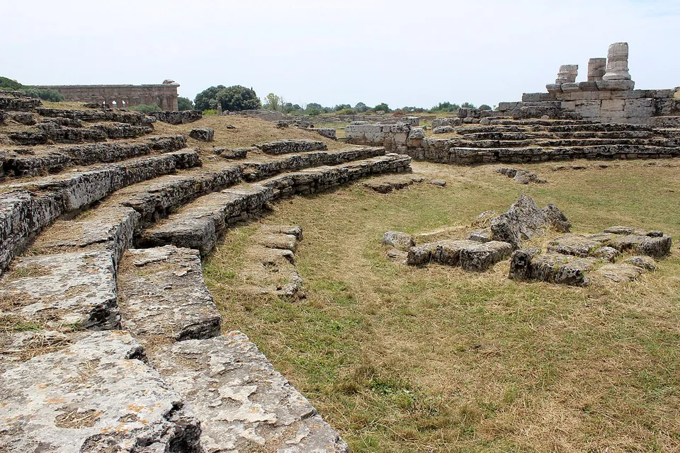 Curving tiers of rough grey stone steps descending into a grassy hollow, with a temple colonnade visible on the horizon.