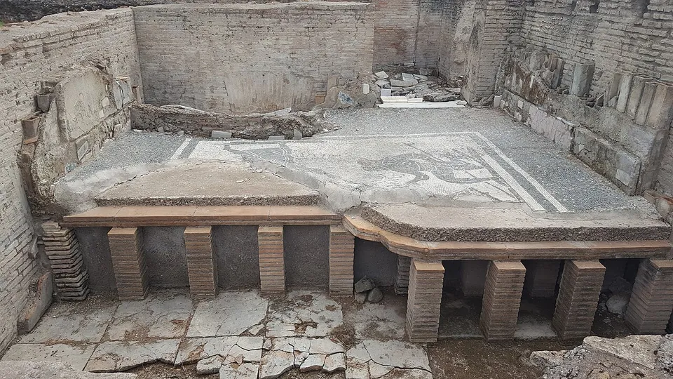 An excavated room in which a black-and-white mosaic floor, partly broken away at the front, rests on rows of low brick pillars, the space beneath it open, with brick and stone walls around.