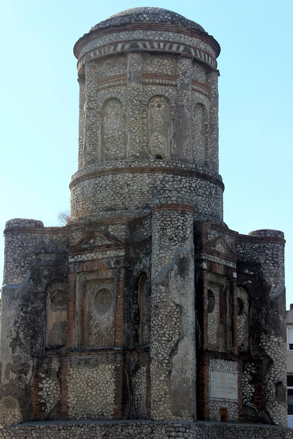 The tomb seen close up from below. A square lower storey with a rounded turret at each corner and recessed panels and niches in its faces carries a round upper drum ringed with arched niches and a low domed top; the walls are faced with small grey stones and edged with red brick.