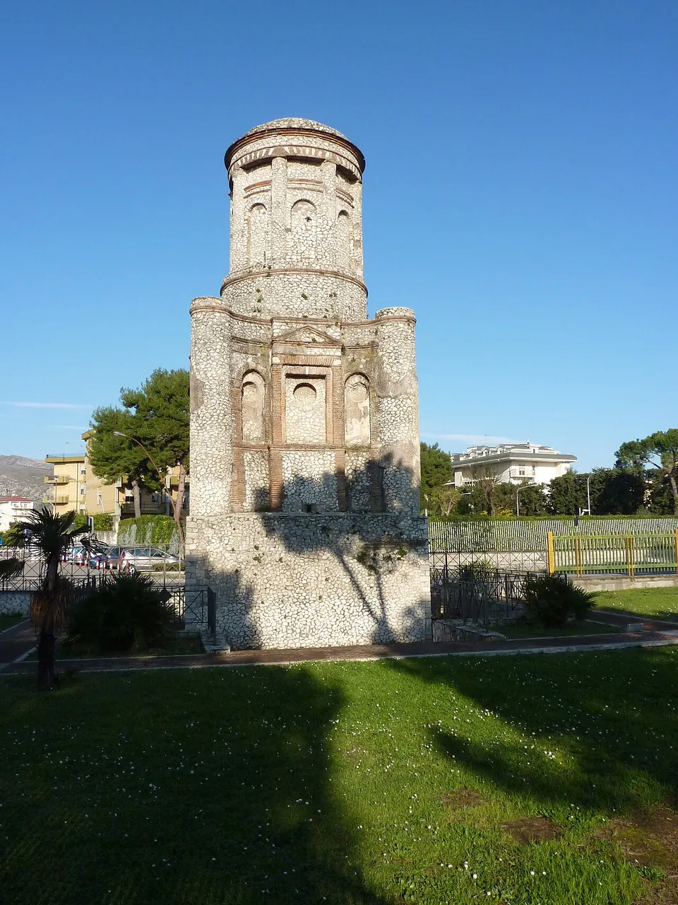The tomb seen across a mown lawn in low sunlight, casting a long shadow towards the camera, with a palm tree and modern buildings behind it.