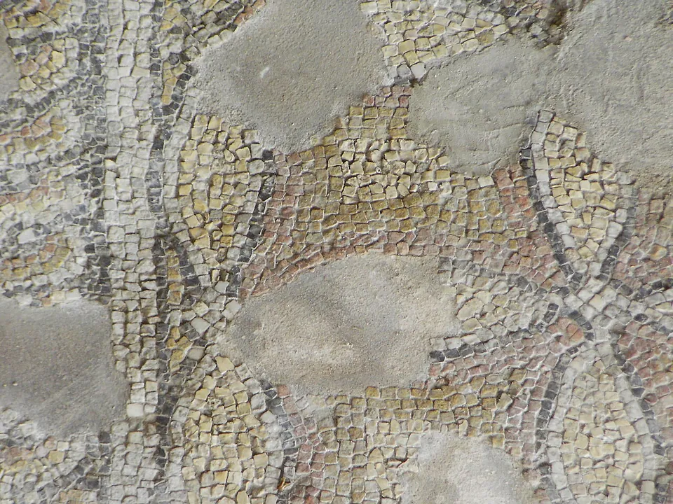 Close view of a mosaic of small stone cubes in cream, ochre and grey, forming curving leaf and scroll shapes, with patches of plain repair.