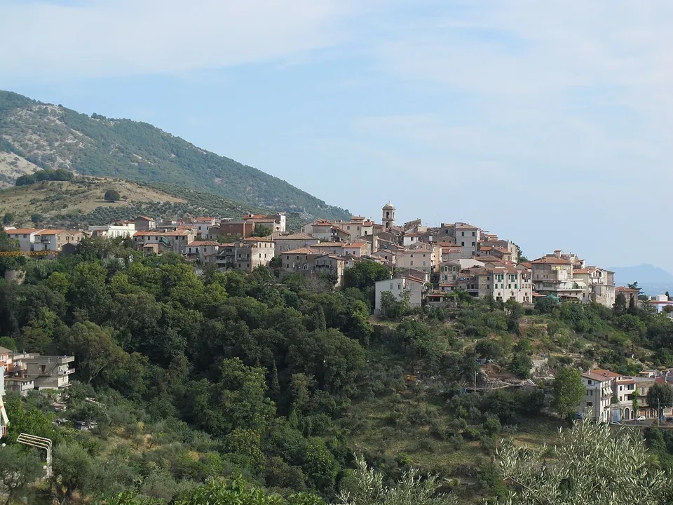 A hill town of pale stone houses spread along a ridge above wooded slopes, with a bell tower among the roofs and a green mountainside rising behind.