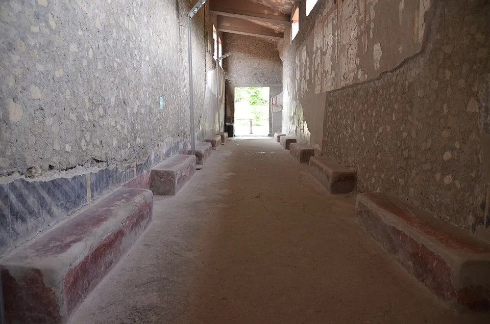 A long, straight, roofed corridor with plastered stone walls, low masonry benches painted red along both sides and bright daylight at the far end.