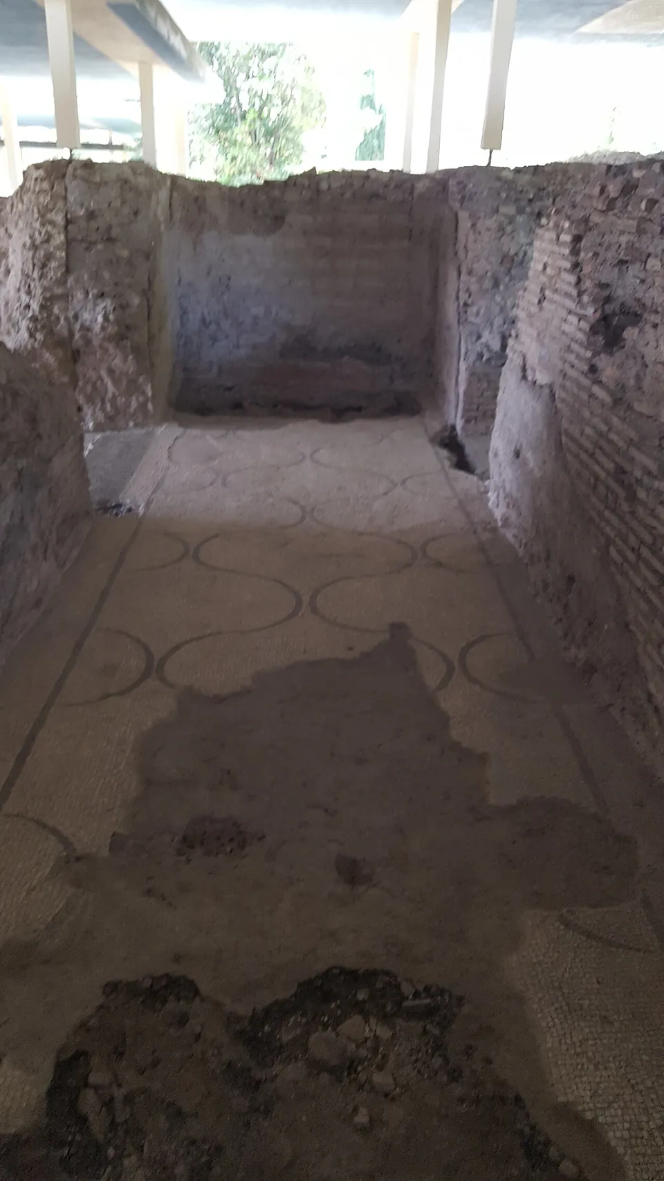 A narrow corridor between brick and rubble walls, its floor a white mosaic with a pattern of interlocking black circles, broken away in patches towards the near end.