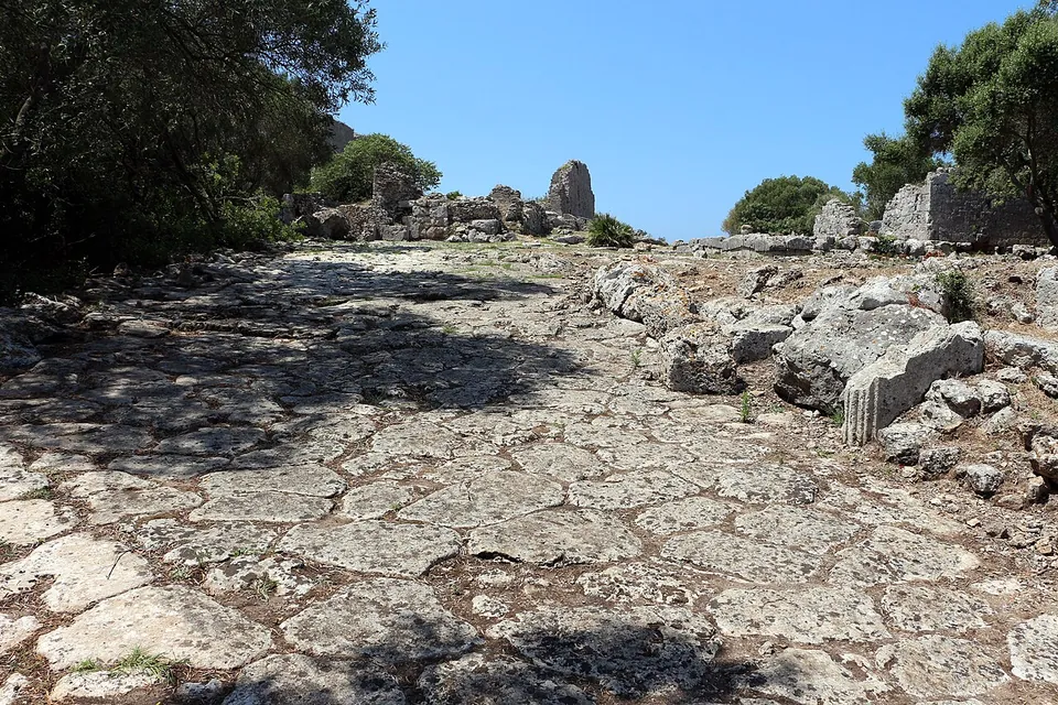 A street surface of large irregular polygonal paving stones climbs gently between scattered fallen blocks, with low ruins and trees at the top of the rise.