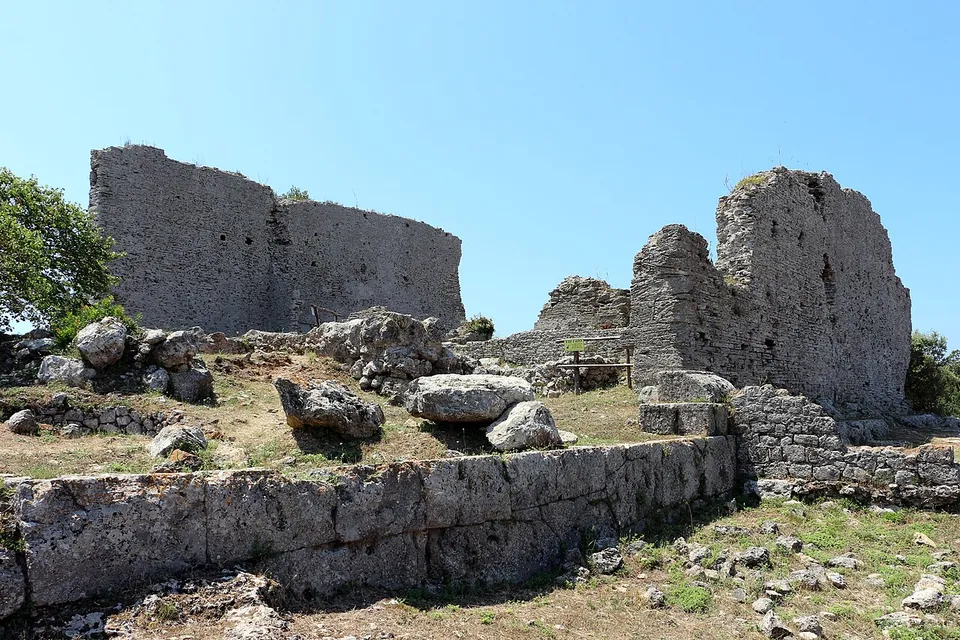 Two tall ruined walls of grey rubble masonry rise above a terrace edged with a course of large squared blocks, with fallen stones scattered in the grass in front and a clear blue sky behind.
