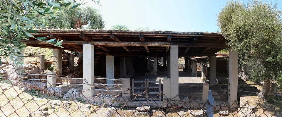A low timber roof on pale square pillars shelters excavated rooms and low walls, seen through a wire fence among olive trees.