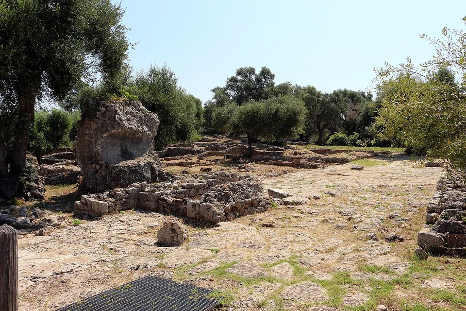A broad open area of bare rock and dry grass with low walls and a broken block of masonry on the left, ringed by olive trees.