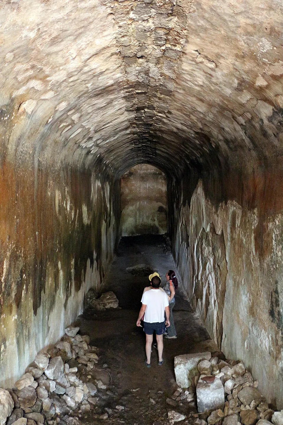 A long barrel-vaulted tunnel with plastered, water-stained walls, two visitors walking along its floor towards the light at the far end.