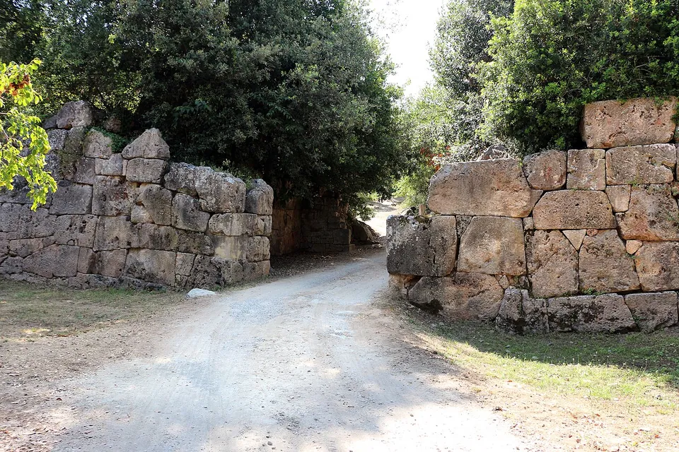 A gravel track runs through a gap in a wall of huge irregular stone blocks fitted without mortar, with trees growing over the wall on both sides.